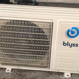 AS - CLIMATECH Arkadiusz Sosna - Biała jednostka zewnętrzna klimatyzacji Blyss, widoczna kratka wentylacyjna i logo firmy, ustawiona na betonowej posadzce.