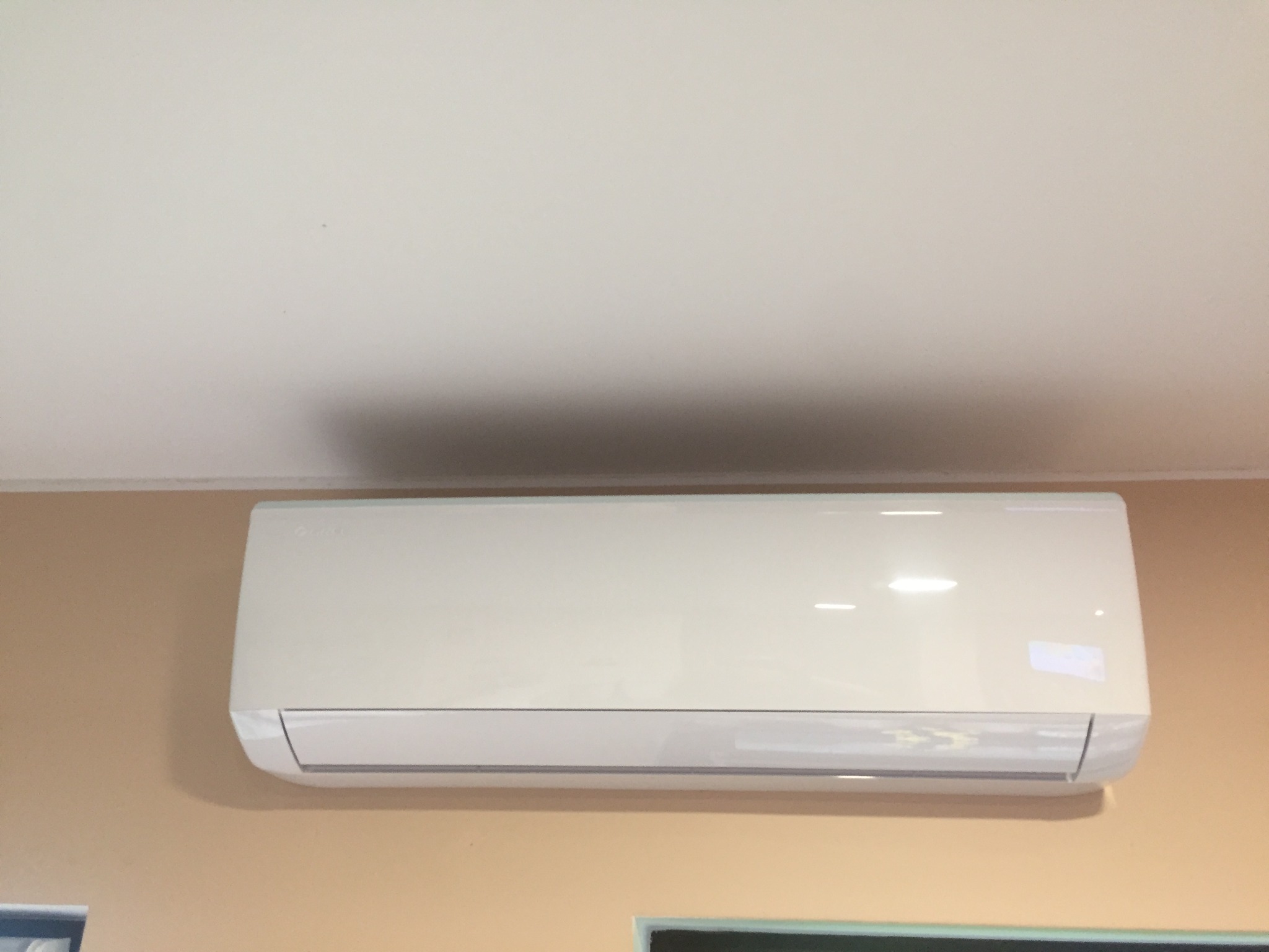 Biała klimatyzacja split na beżowej ścianie pod białym sufitem. Widoczne okno w dolnej części kadru. Oświetlenie dzienne.