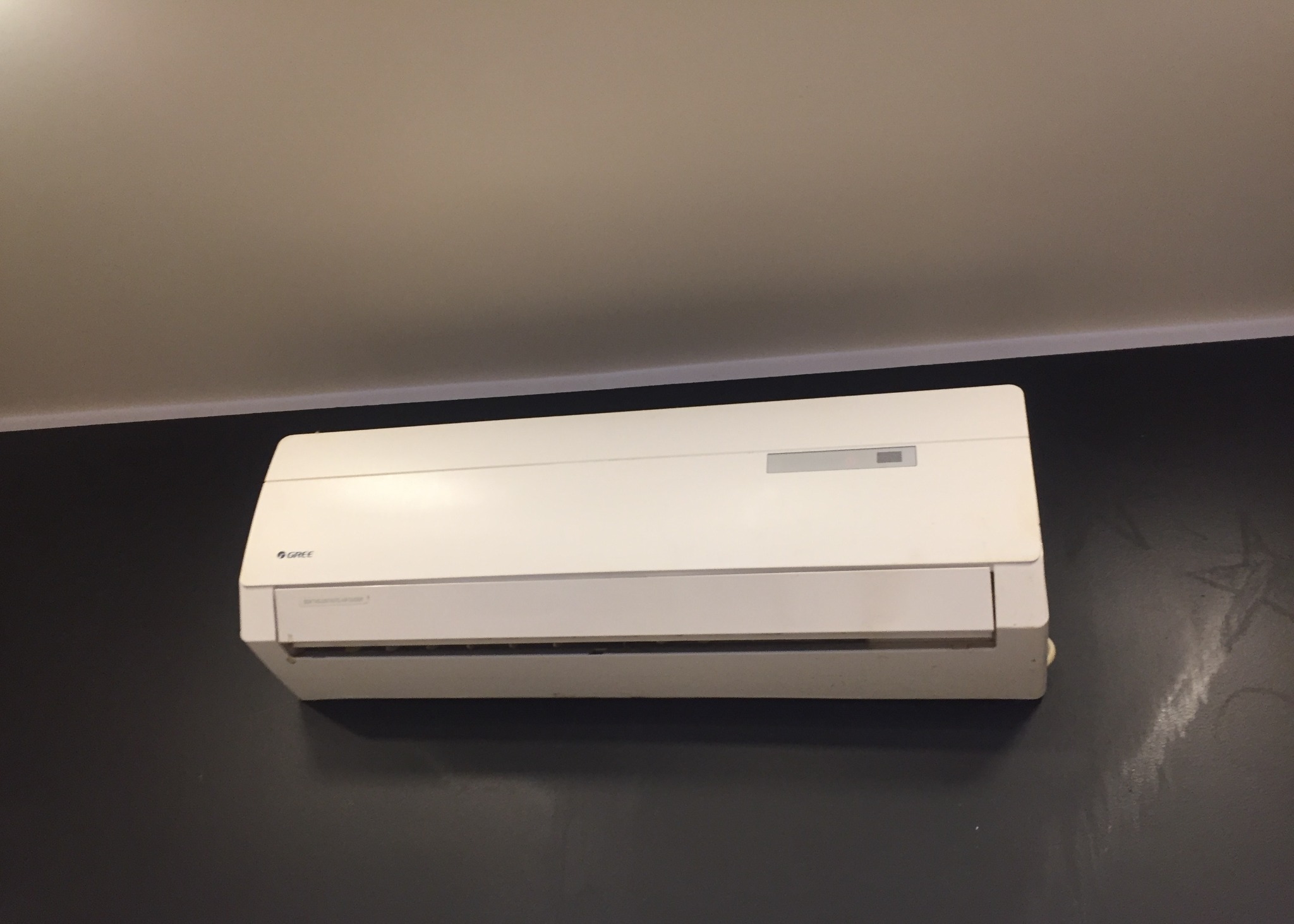 Biała klimatyzacja marki Gree, zamontowana na ciemnej ścianie pod jasnym sufitem. Widoczne detale urządzenia i otoczenia.