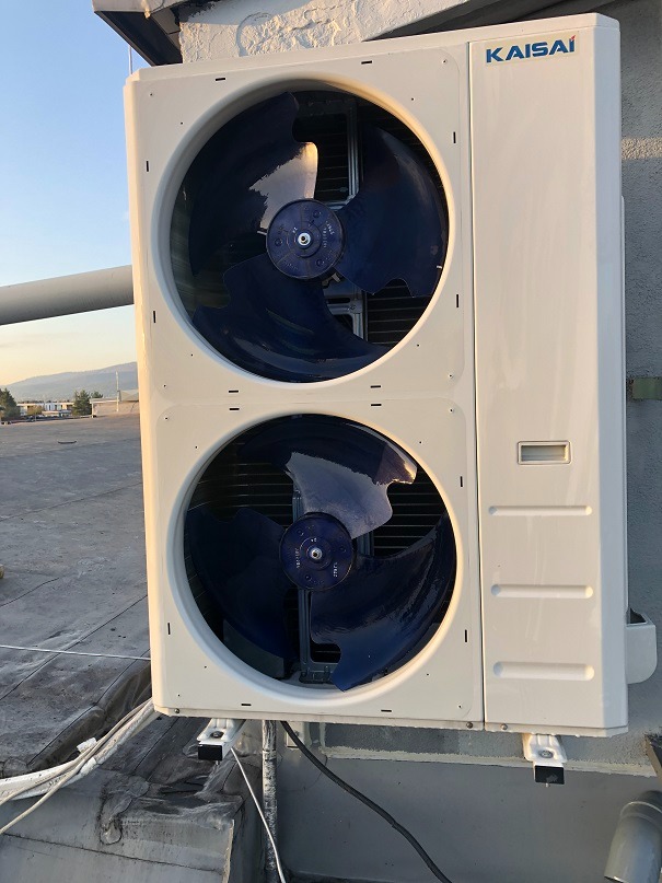 Biała, dwu-wentylatorowa jednostka zewnętrzna klimatyzacji Kaisai zamontowana na dachu budynku, z widocznymi przewodami. W tle krajobraz miejski o zmierzchu.