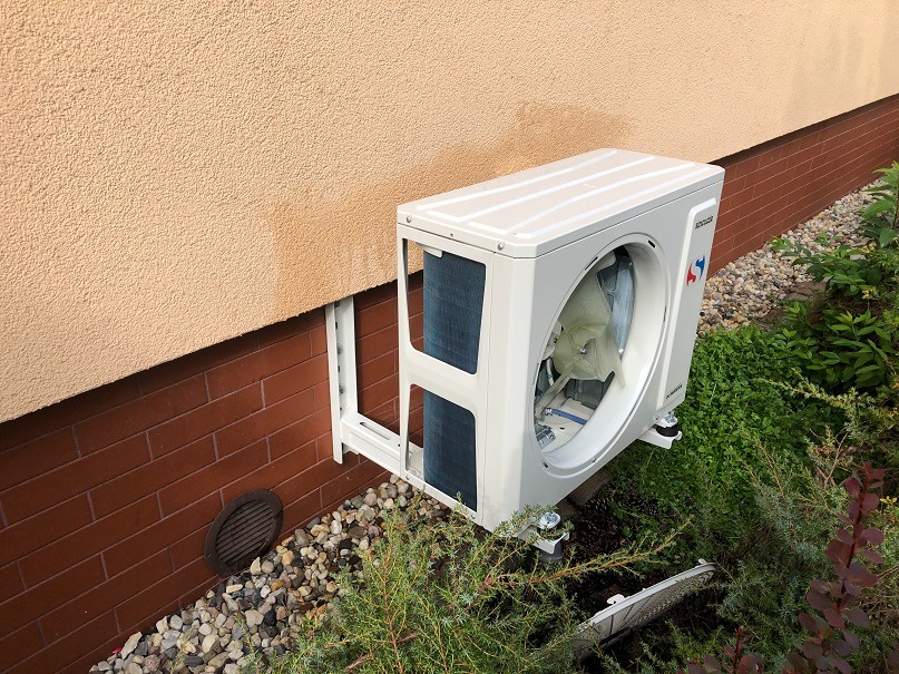 Demontaż klimatyzatora zewnętrznego na ścianie budynku z widoczną instalacją i elementami montażowymi. Widoczne rośliny w tle.