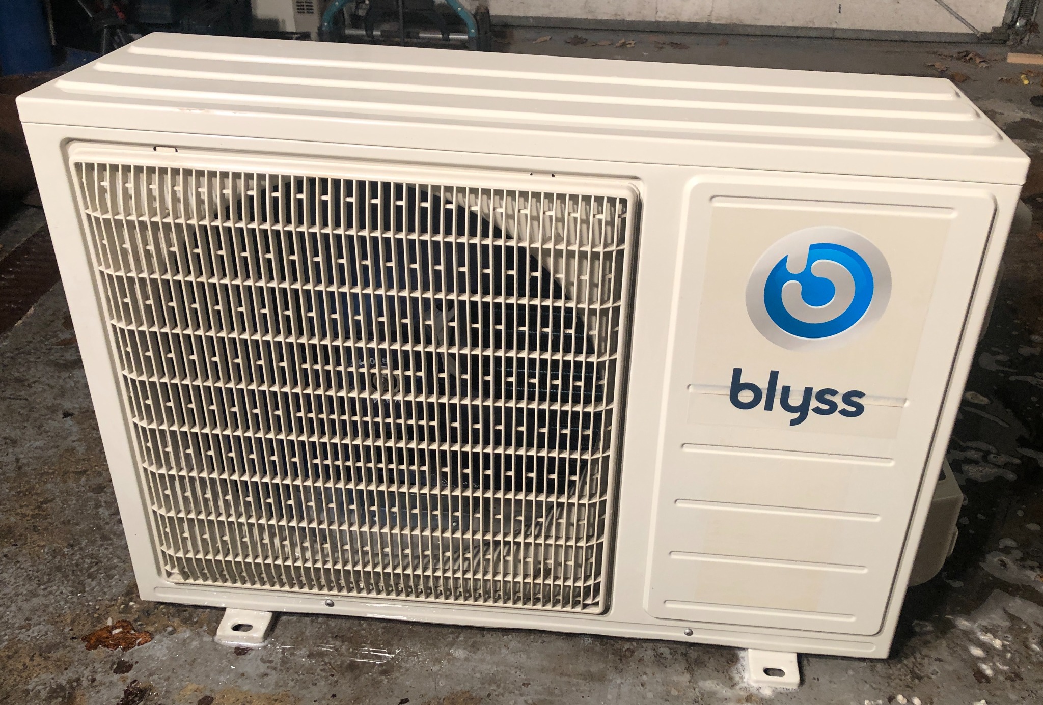 Biała jednostka zewnętrzna klimatyzacji Blyss, widoczna kratka wentylacyjna i logo firmy, ustawiona na betonowej posadzce.