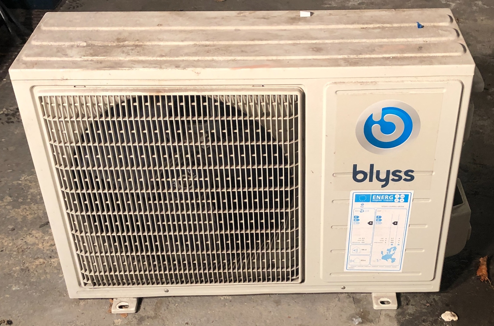 Blyss - zewnętrzna jednostka klimatyzacji z widoczną kratką wentylacyjną i etykietą energetyczną, lekko przybrudzona, stojąca na betonowym podłożu.