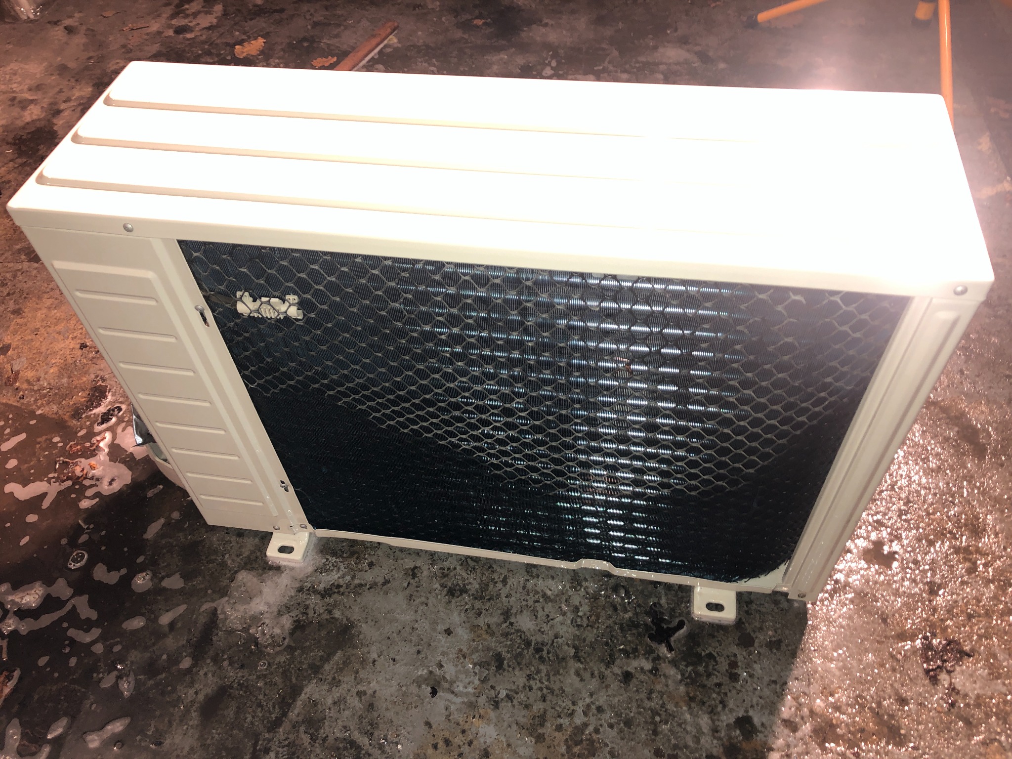 Klimatyzator zewnętrzny, widok z góry pod kątem, ukazujący metalową siatkę ochronną i beżową obudowę na tle wilgotnej posadzki.