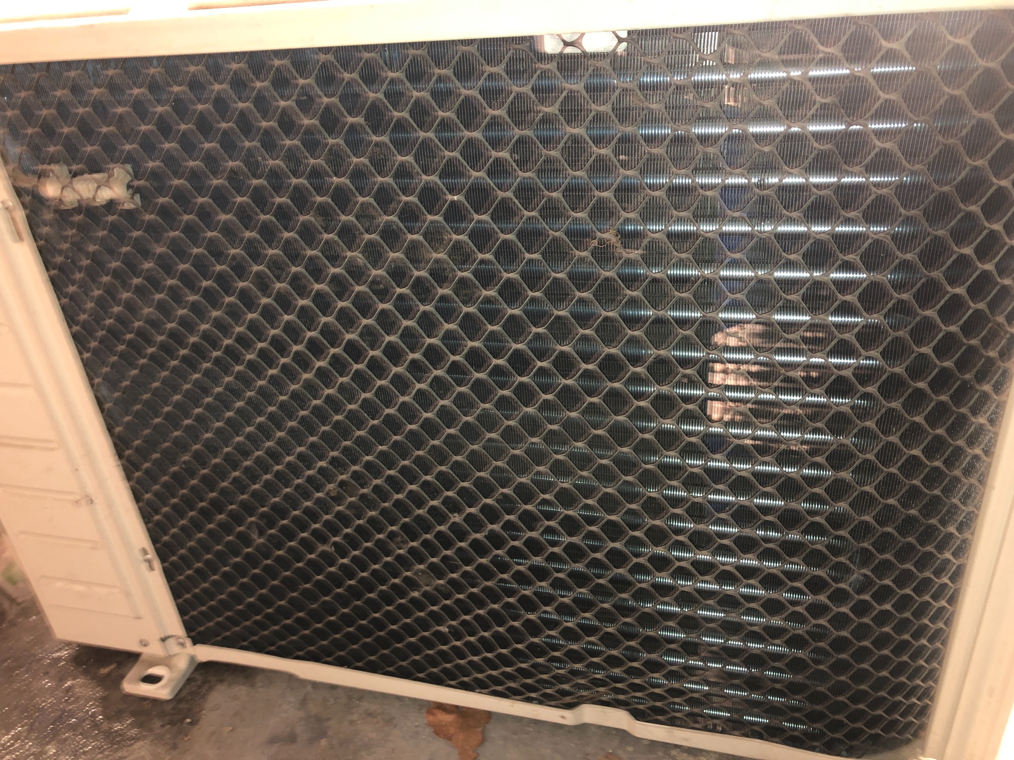 Zanieczyszczona kratka wentylacyjna klimatyzatora, widoczne drobinki kurzu i brudu na powierzchni. Metalowa konstrukcja z regularnymi otworami, jasne tło wewnątrz.