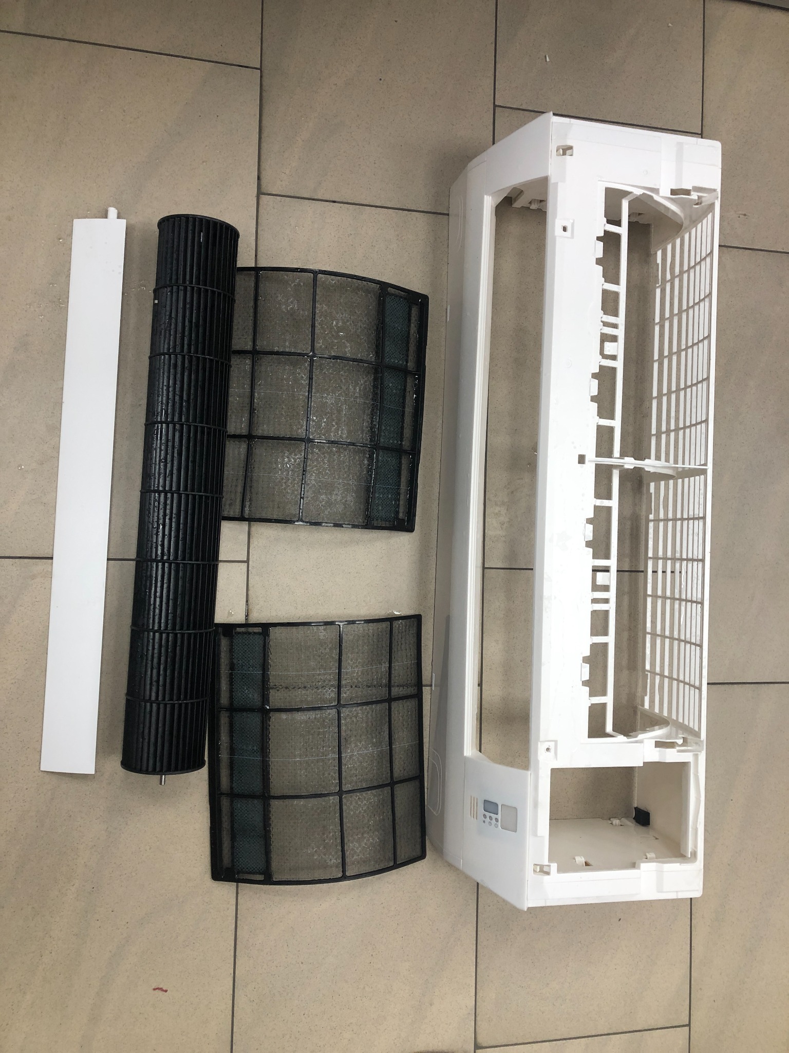 Rozłożony na części klimatyzator: obudowa, filtr siatkowy i filtr walcowy leżą na kafelkach. Widok z góry, ukazujący elementy po demontażu, prawdopodobnie w trakcie czyszczenia.