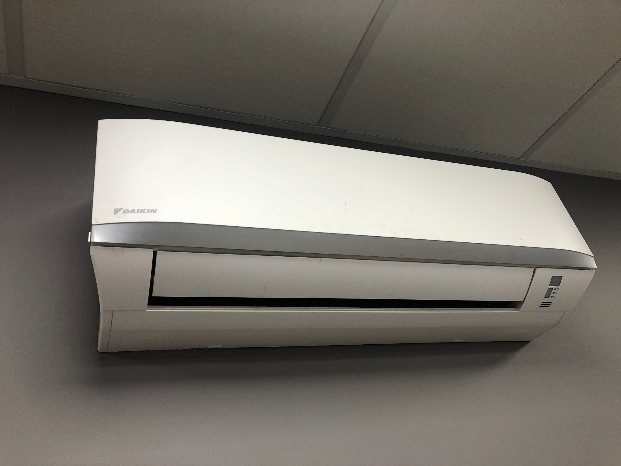 Biała klimatyzacja Daikin zamontowana na szarej ścianie. Widoczny fragment sufitu. Urządzenie skierowane w lewo, z widocznymi detalami obudowy.