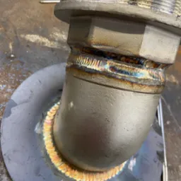 Szczegółowe zbliżenie na metalowy element rurowy po spawaniu, z widocznymi kolorowymi śladami ciepła i precyzyjnymi spoinami. Widoczna faktura metalu i ślady obróbki.