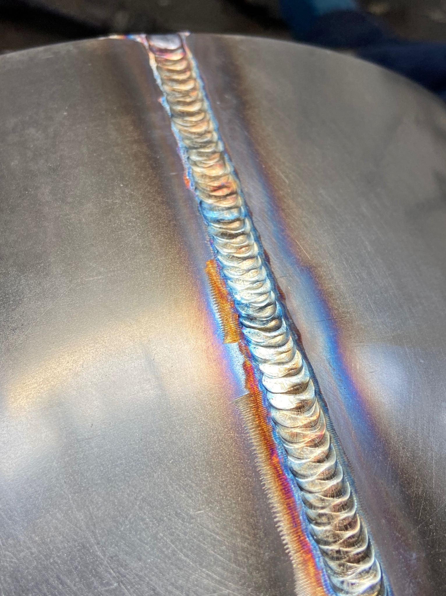 Błyszczące, metalowe złącze spawane z efektem tęczy na krawędziach, ukazujące precyzję i estetykę wykonania. Detal spoiny na gładkiej powierzchni.