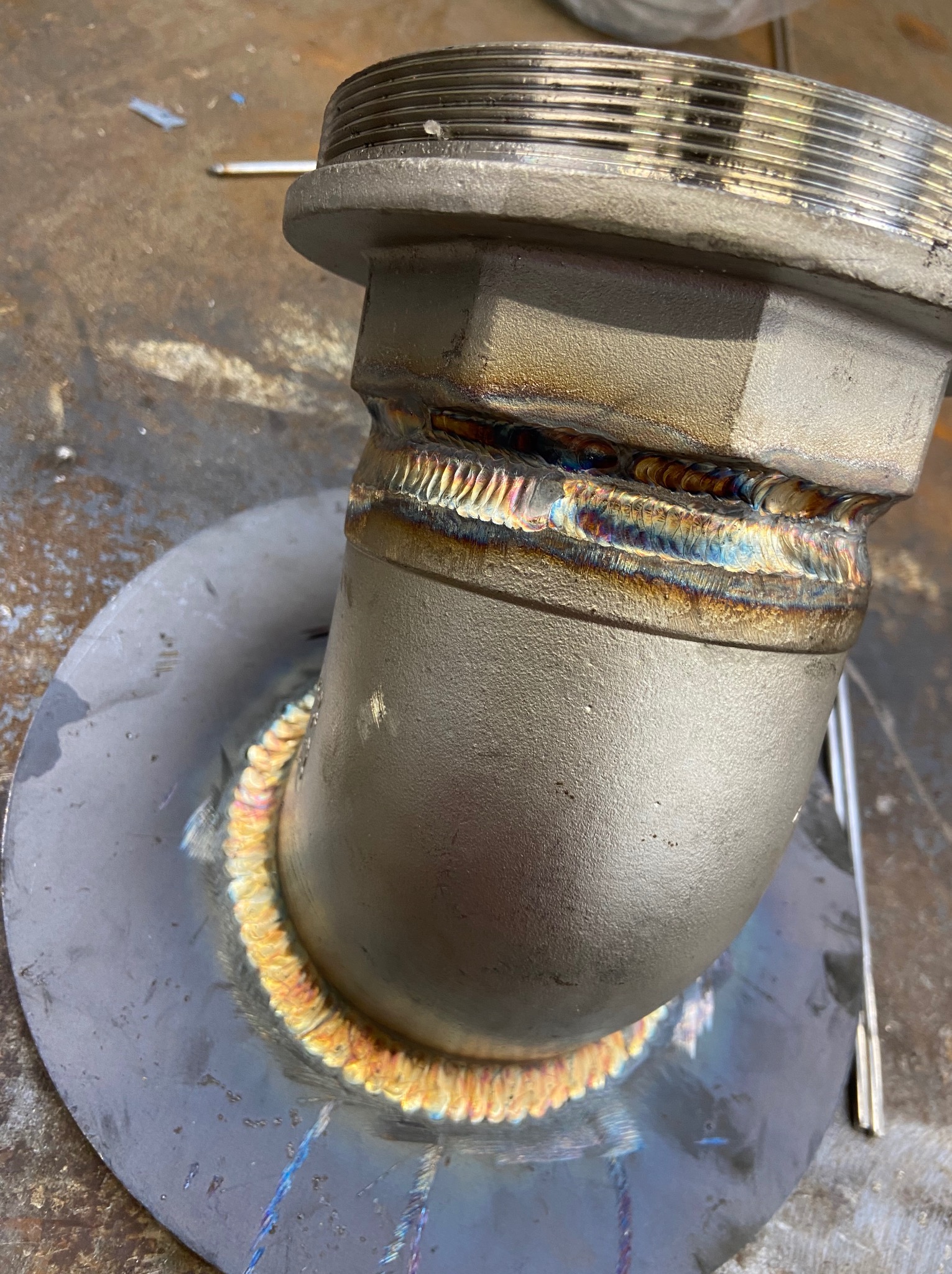 Szczegółowe zbliżenie na metalowy element rurowy po spawaniu, z widocznymi kolorowymi śladami ciepła i precyzyjnymi spoinami. Widoczna faktura metalu i ślady obróbki.