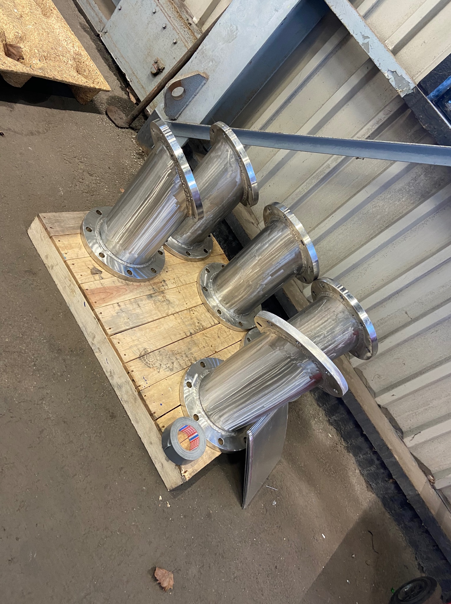 Cztery spawane, nierdzewne rury z kołnierzami leżą na drewnianej palecie w warsztacie. Widoczna taśma klejąca i blaszane elementy.
