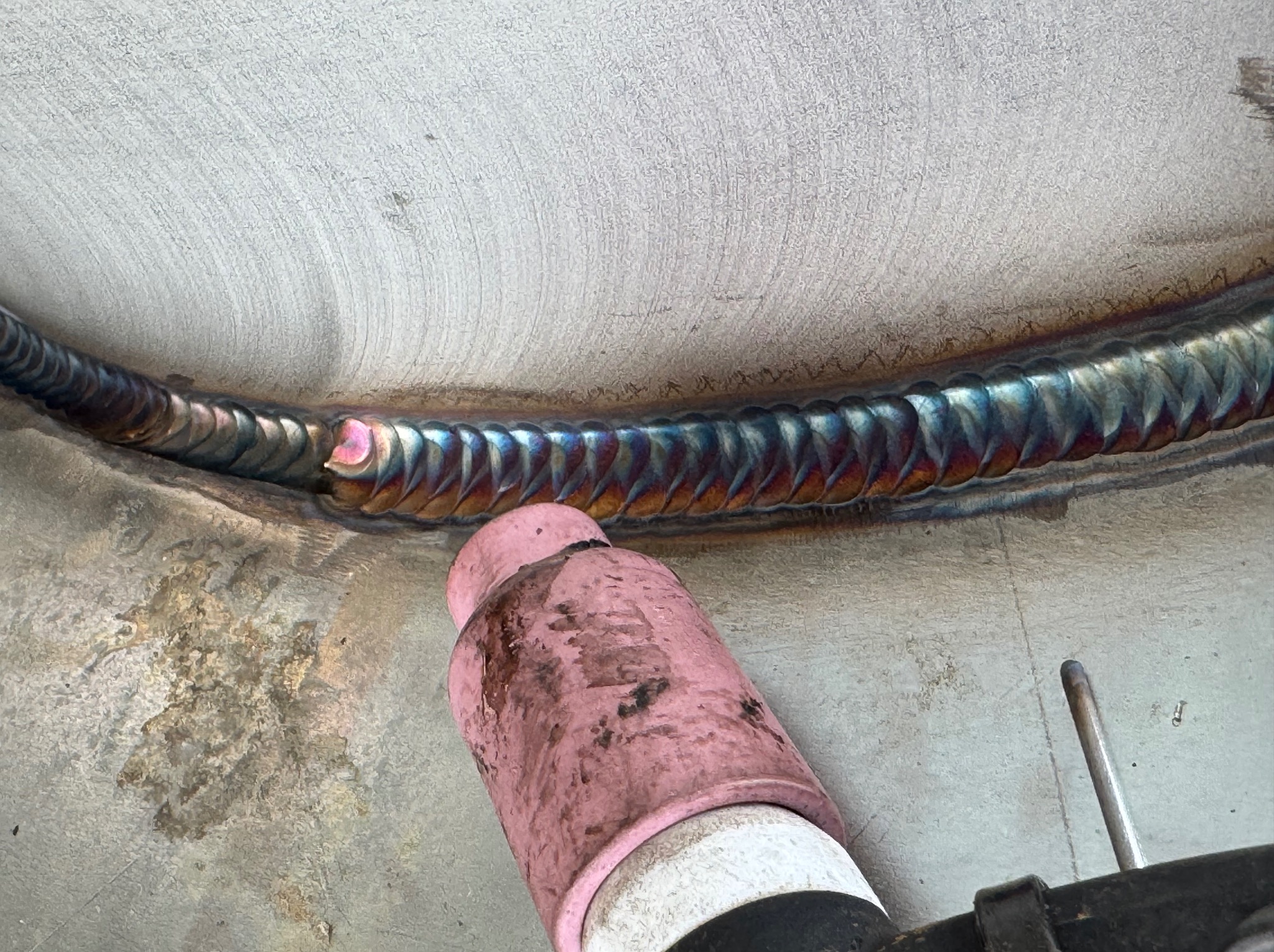 Metalowa powierzchnia po spawaniu metodą TIG z widocznym palnikiem i spoiną o charakterystycznych kolorach. Detal pracy spawacza, zbliżenie na spoinę.