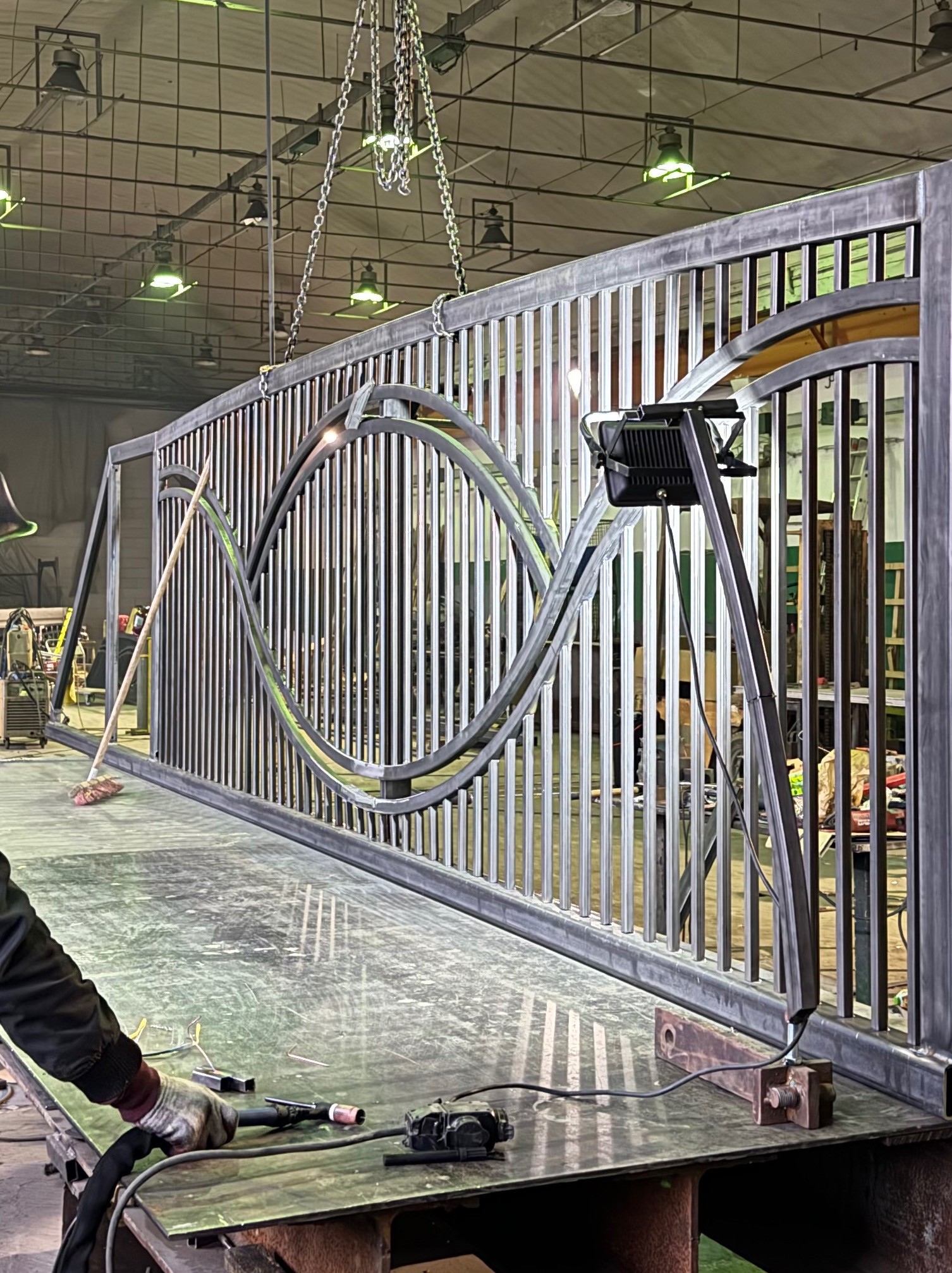 Wytwarzanie metalowej bramy z dekoracyjnymi elementami w warsztacie. Widoczny spawacz przy pracy, lampa oświetleniowa i narzędzia na stole roboczym.