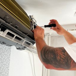 KlimaFM - Serwisant z tatuażem naprawia klimatyzator na ścianie z wzorzystą tapetą. Używa śrubokręta do regulacji wewnętrznych elementów urządzenia.