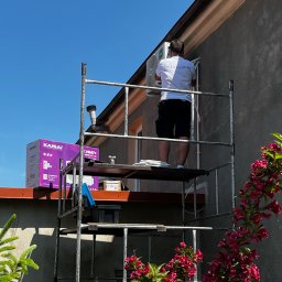 KlimaFM - Montaż klimatyzacji zewnętrznej na rusztowaniu. Widoczna jednostka, instalator w koszulce z logo, pudełko Kaisai i kwitnący krzew na pierwszym planie.