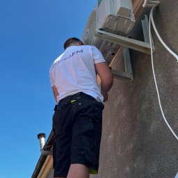 KlimaFM - Instalator montuje jednostkę zewnętrzną klimatyzacji na elewacji budynku, widok z dołu na tle błękitnego nieba. Widoczne rury i przewody instalacyjne.