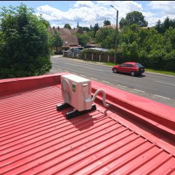 KlimaFM - Biała jednostka zewnętrzna klimatyzacji Kaisai zamontowana na czerwonym blaszanym dachu budynku, widok na ulicę i zieleń w tle.