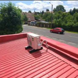 Biała jednostka zewnętrzna klimatyzacji Kaisai zamontowana na czerwonym blaszanym dachu budynku, widok na ulicę i zieleń w tle.