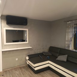 KlimaFM - Nowoczesny klimatyzator Karsai w salonie z szarą ścianą, lustrem w białej ramie i czarno-białą sofą narożną. Widoczny wentylator w rogu pokoju.