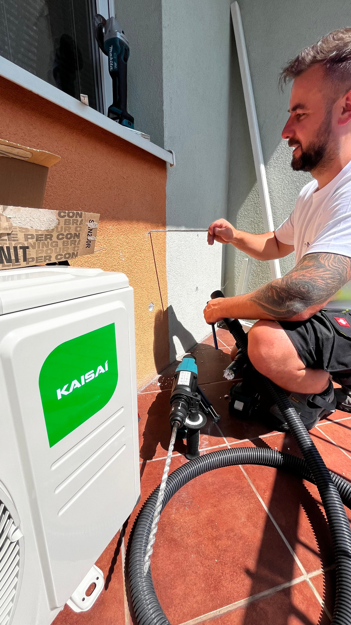 Montaż klimatyzacji Kaisai: instalator wierci otwór w ścianie obok jednostki zewnętrznej. Widoczna wiertarka, odkurzacz i narzędzia na zewnątrz budynku.