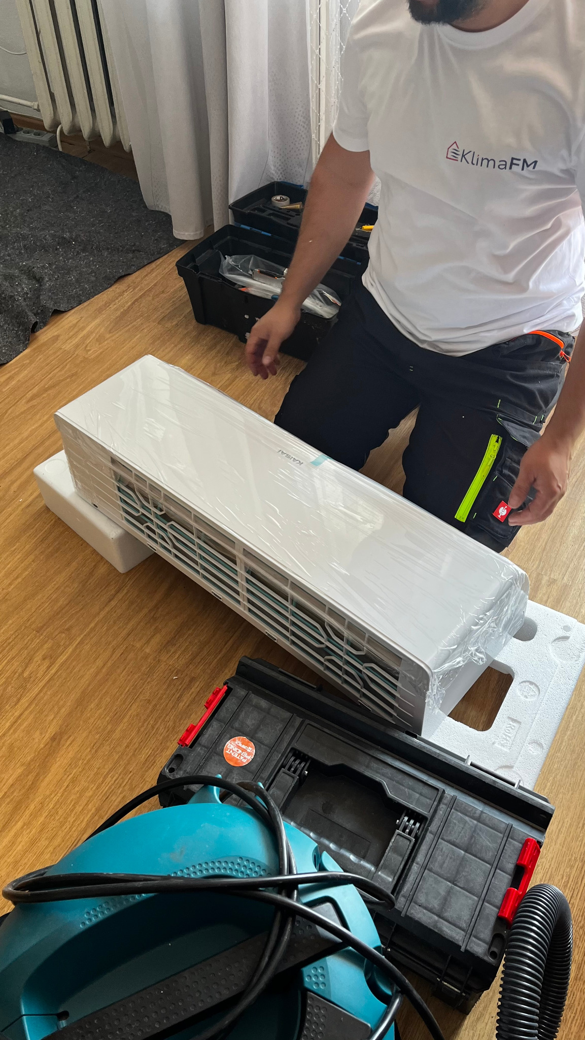 Biała jednostka klimatyzacji Kaisai w folii, leży na podłodze obok narzędzi i skrzynki narzędziowej. Widoczny instalator w koszulce z logo KlimaFM.