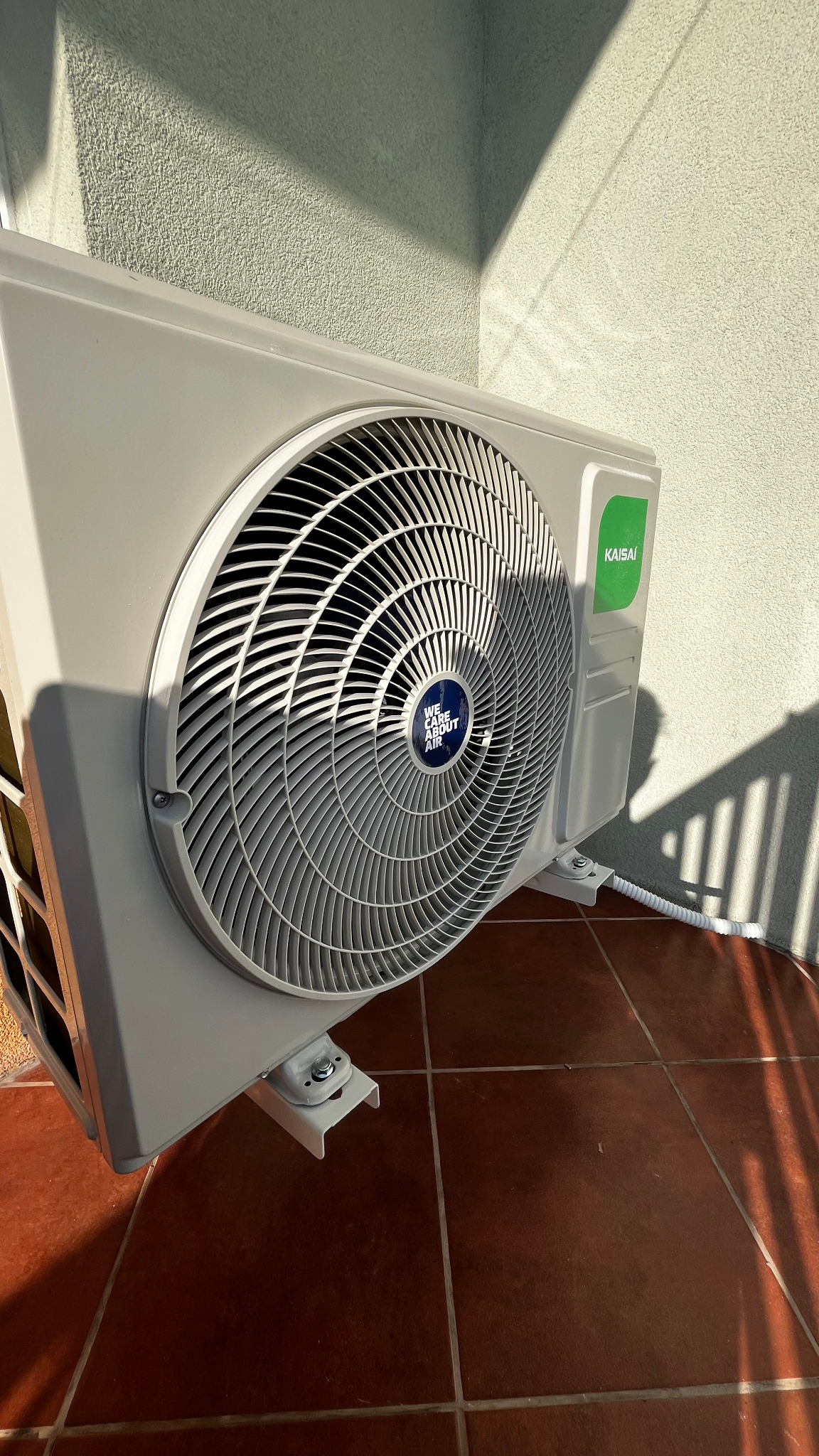 Zamontowana jednostka zewnętrzna klimatyzacji Kaisai na balkonie, widoczne mocowania i przewód odprowadzający skropliny, cień rzucany przez balustradę.