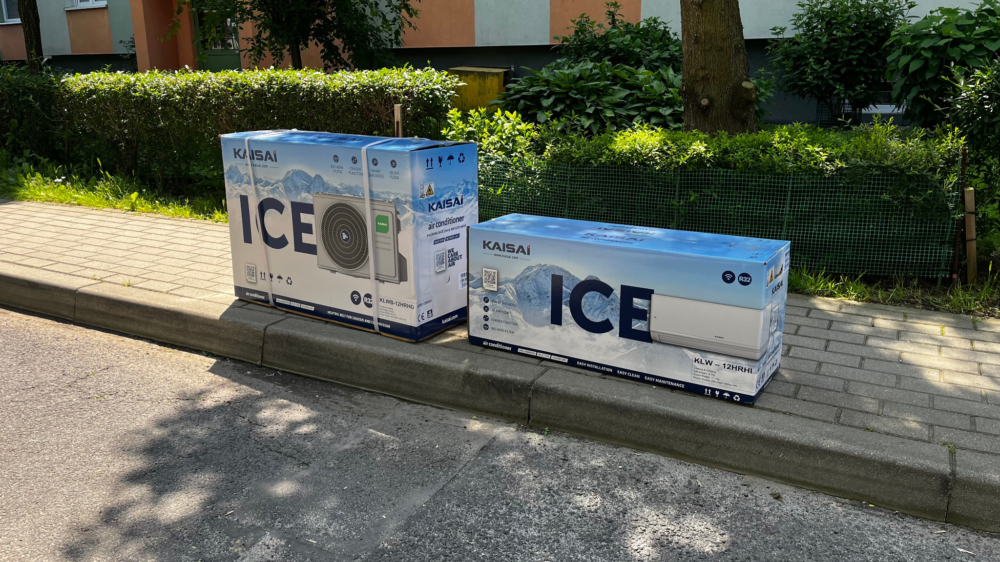Dwa kartony klimatyzatorów Kaisai ICE leżące na chodniku przy krawężniku, na tle zieleni. Widoczne modele KLWB-12HRHO i KLW-12HRHI. Oznakowania: R32, easy installation.
