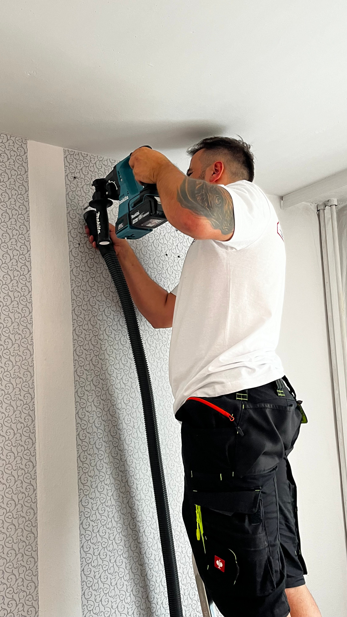 Pracownik z wiertarką Makita i odkurzaczem podczas montażu wentylacji w Lubaniu. Widoczne otwory w ścianie z tapetą w dekoracyjne wzory.
