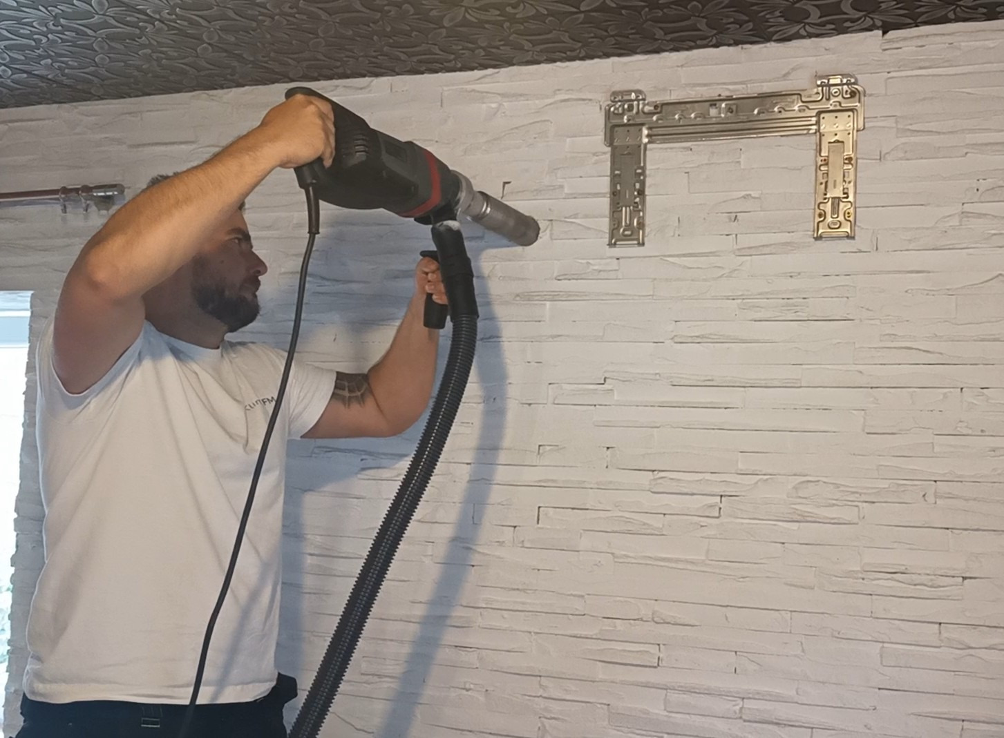 Mężczyzna wierci otwór w ścianie z dekoracyjnej cegły, używając wiertarki z odkurzaczem, obok zamontowany uchwyt klimatyzacji.