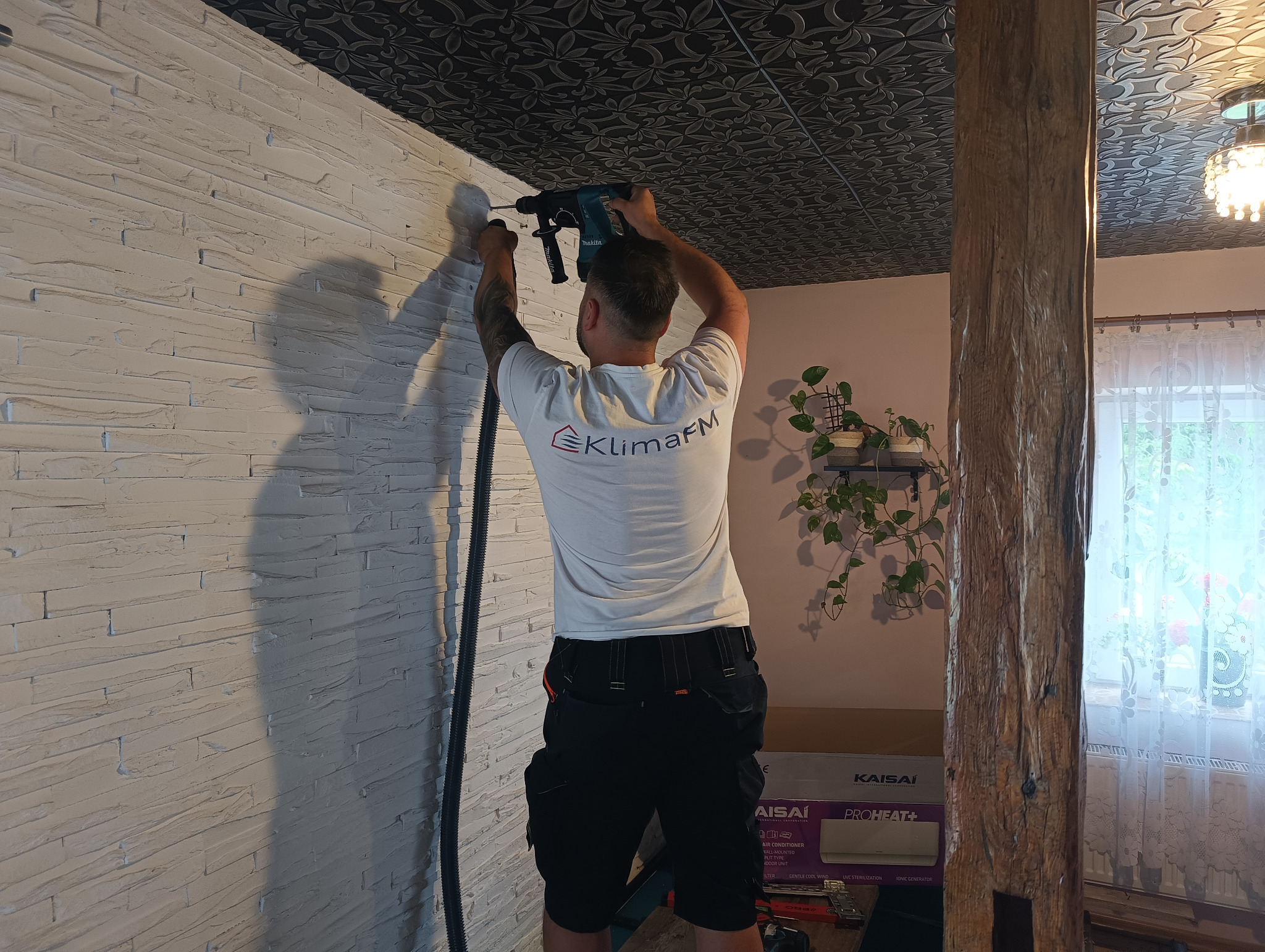 Instalator wierci otwór w ścianie z cegły, przygotowując miejsce na klimatyzację. Widoczny sprzęt i logo firmy KlimaFM na koszulce. W tle sufit z wzorem.