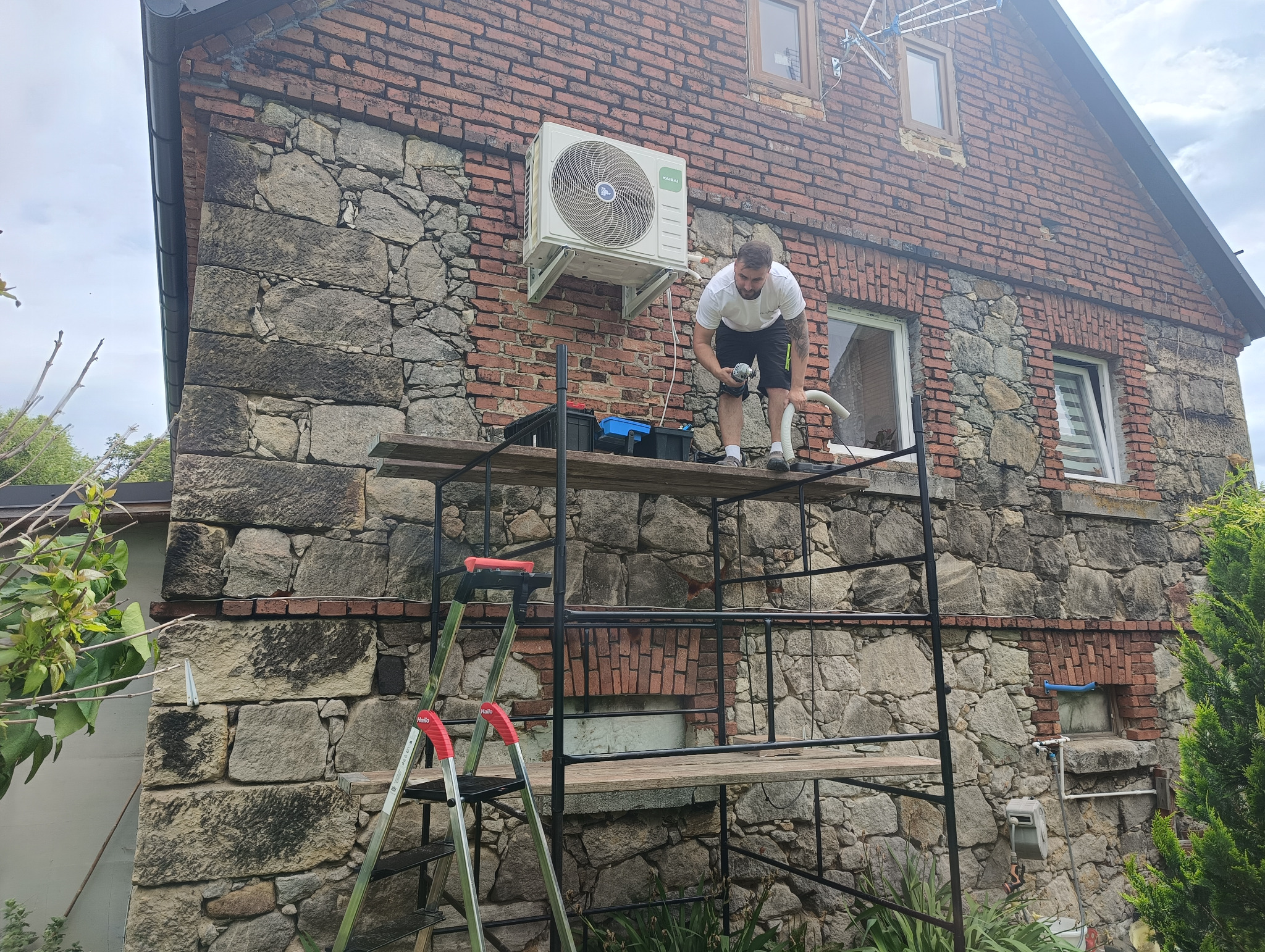 Montaż klimatyzacji na elewacji z kamienia i cegły. Mężczyzna na rusztowaniu instaluje klimatyzator marki Kaisai. Widoczna drabina i narzędzia.