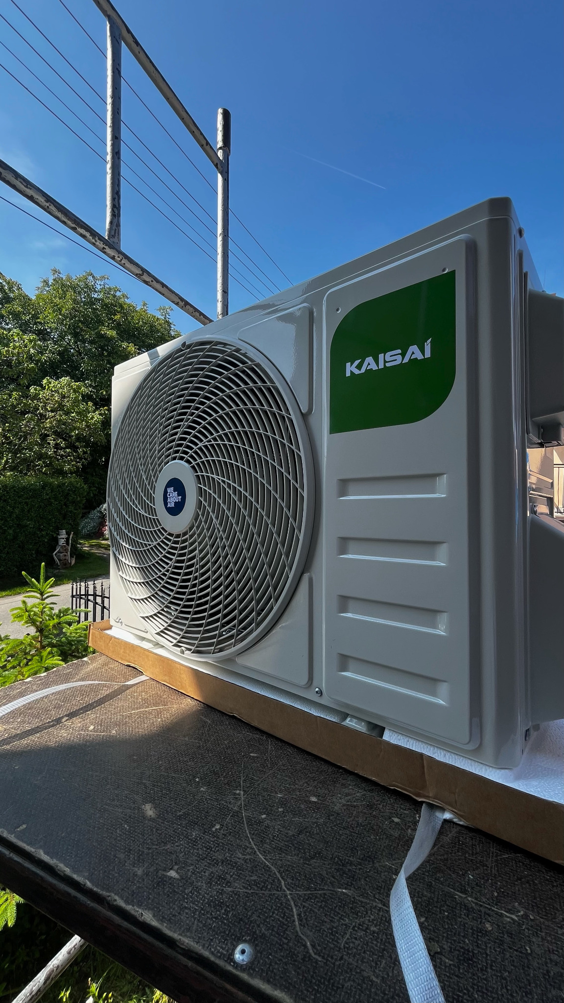 Kompaktowa jednostka zewnętrzna klimatyzacji Kaisai, widoczna z boku, z zielonym logo na tle błękitnego nieba i fragmentu rusztowania.