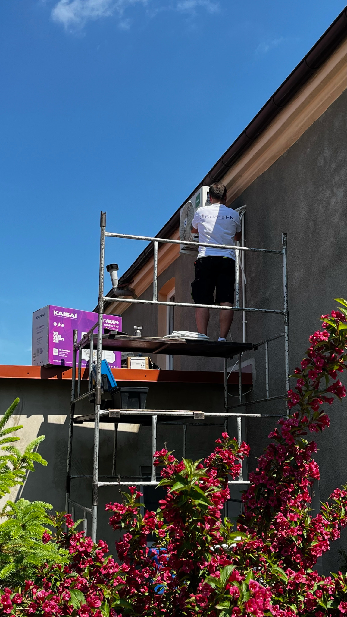 Montaż klimatyzacji zewnętrznej na rusztowaniu. Widoczna jednostka, instalator w koszulce z logo, pudełko Kaisai i kwitnący krzew na pierwszym planie.