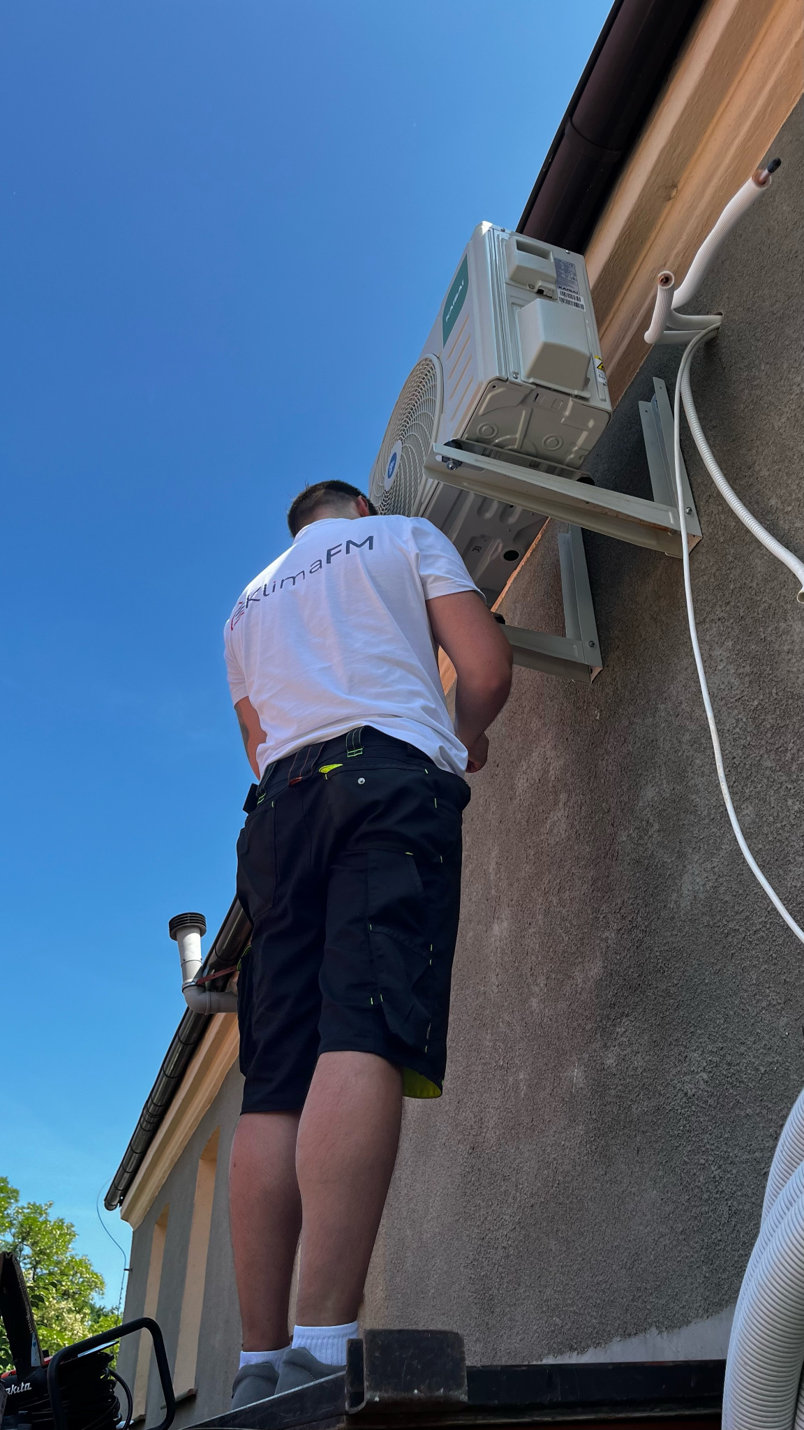 Instalator montuje jednostkę zewnętrzną klimatyzacji na elewacji budynku, widok z dołu na tle błękitnego nieba. Widoczne rury i przewody instalacyjne.