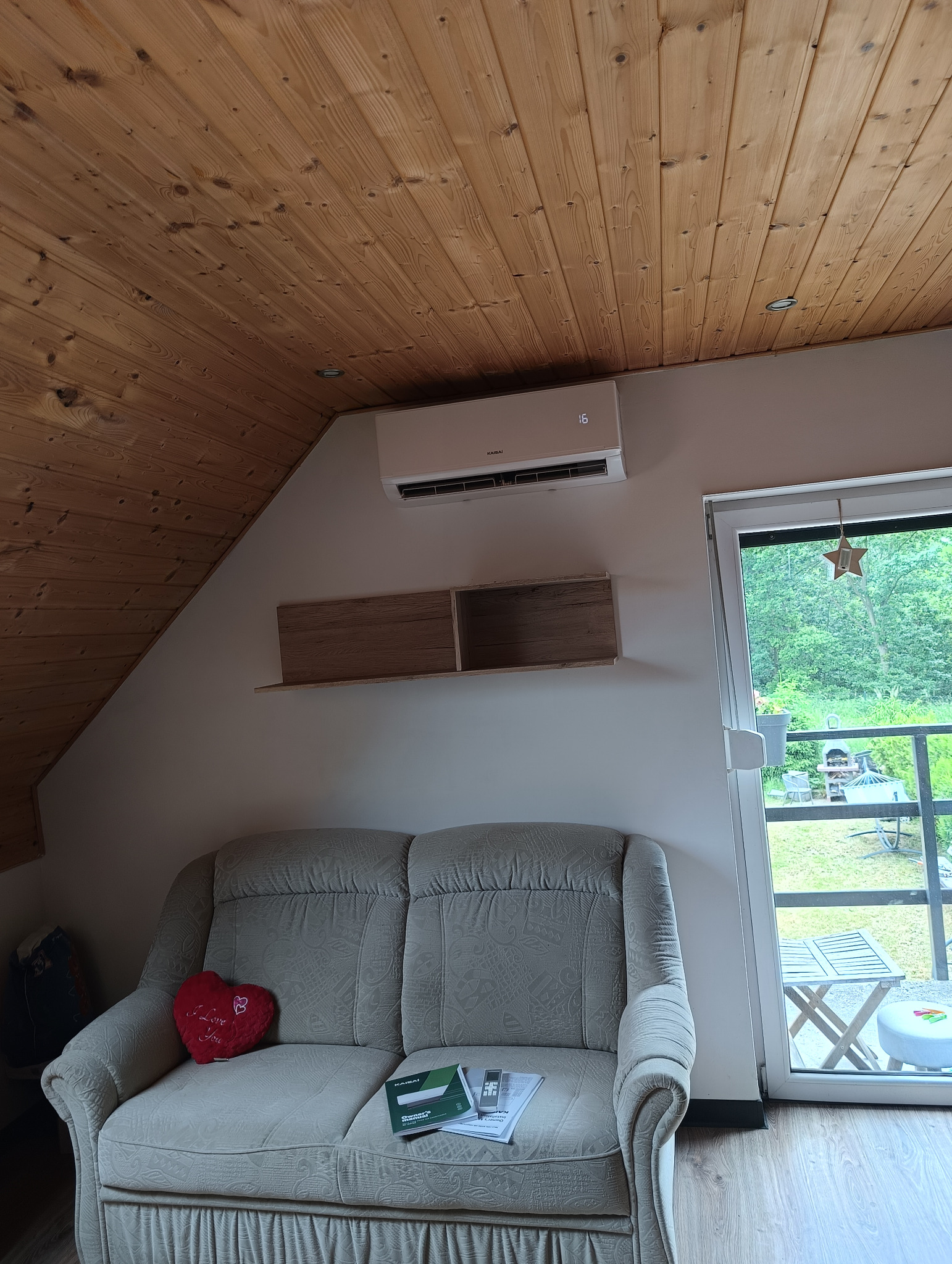 Klimatyzator Kaisai w jasnym wnętrzu z drewnianym stropem. Na sofie leżą instrukcje i pilot do klimatyzacji. Widok na zewnątrz przez drzwi balkonowe.