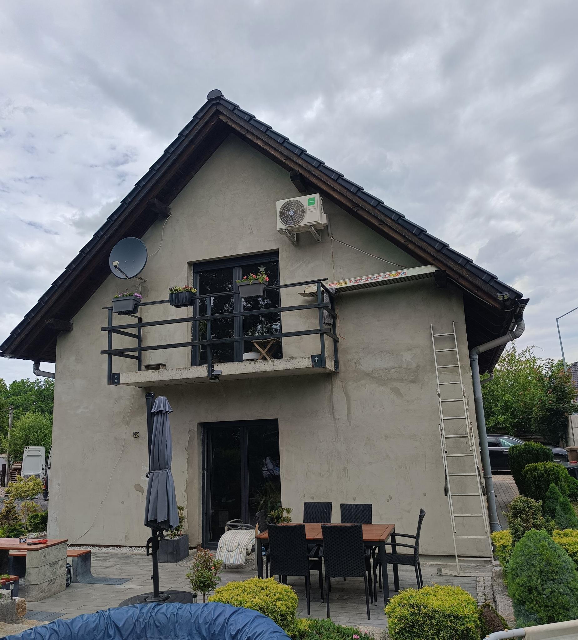 Montaż klimatyzacji na elewacji domu z balkonem. Widoczna drabina oparta o ścianę, stół z krzesłami, satelita i kwiaty w donicach na balkonie.