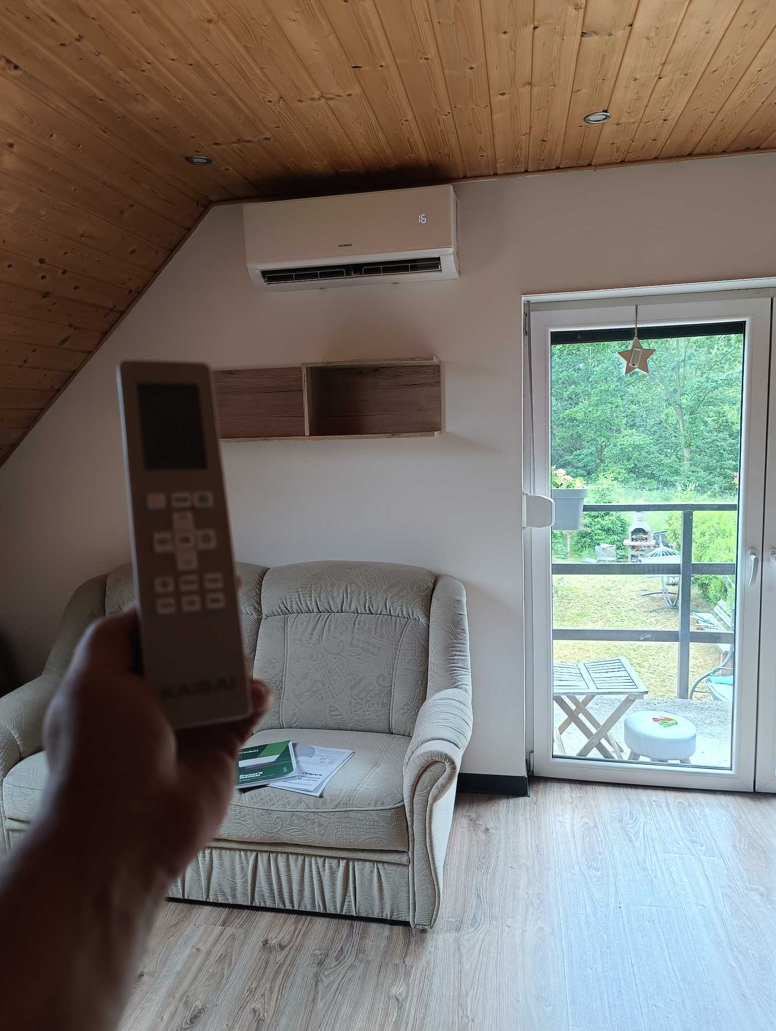 Klimatyzator Kaisaia z pilotem w dłoni, zamontowany w jasnym wnętrzu z drewnianym sufitem i widokiem na zieleń przez okno.