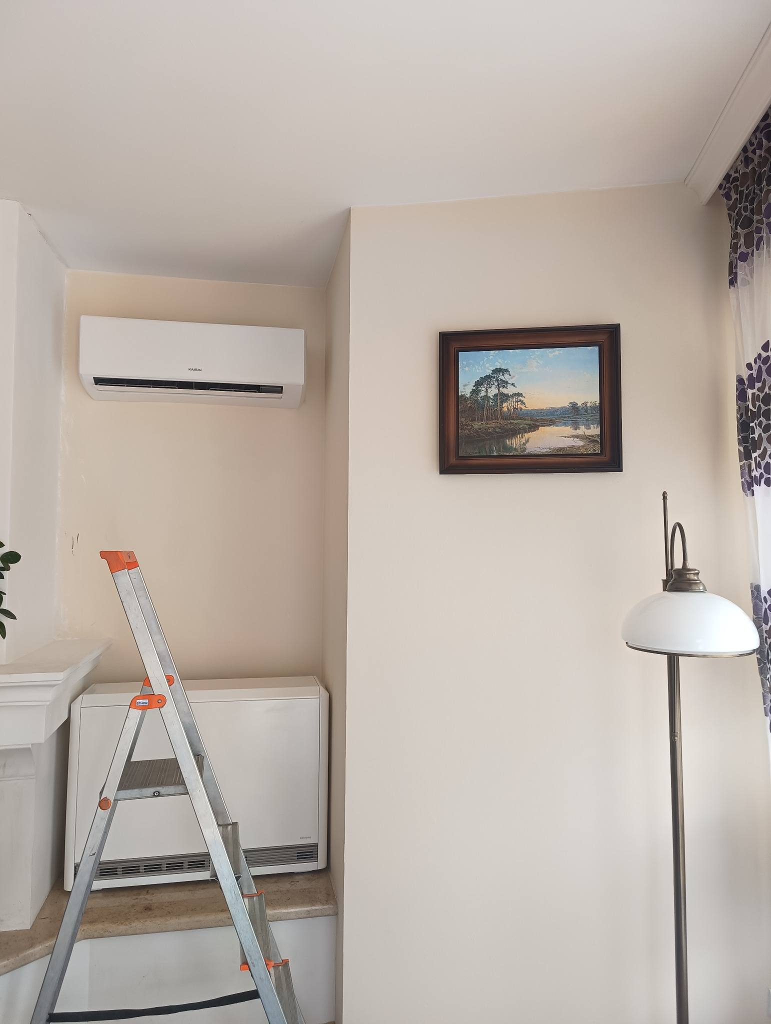 Biały klimatyzator Kaisai zamontowany na ścianie w jasnym pokoju, widoczna drabina i grzejnik. Obrazek na ścianie i lampa dodają wnętrzu charakteru.