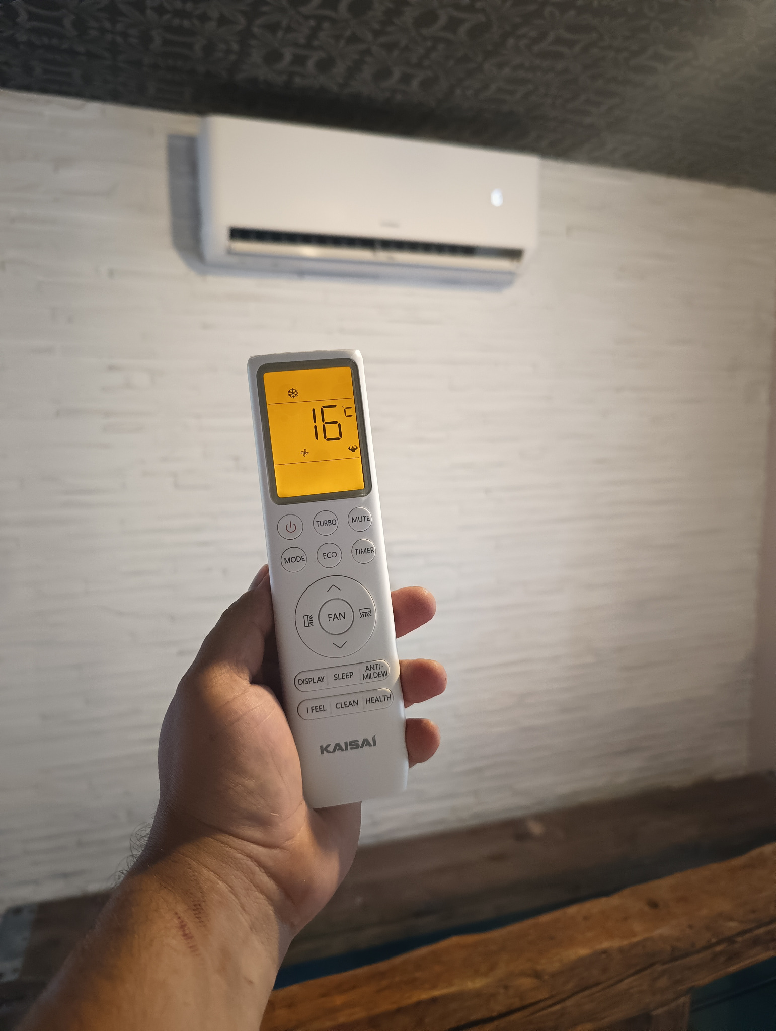 Biały klimatyzator na ścianie, sterowany pilotem marki Kaisai ustawionym na 16 stopni Celsjusza. Widoczna dłoń trzymająca pilot.