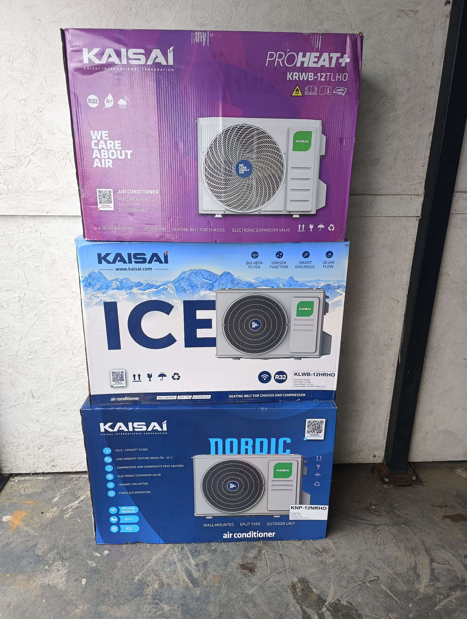 Stos pudełek klimatyzatorów Kaisai: ProHeat+, Ice i Nordic. Etykiety wskazują na modele ścienne, split i zewnętrzne. Widoczne specyfikacje techniczne i cechy.