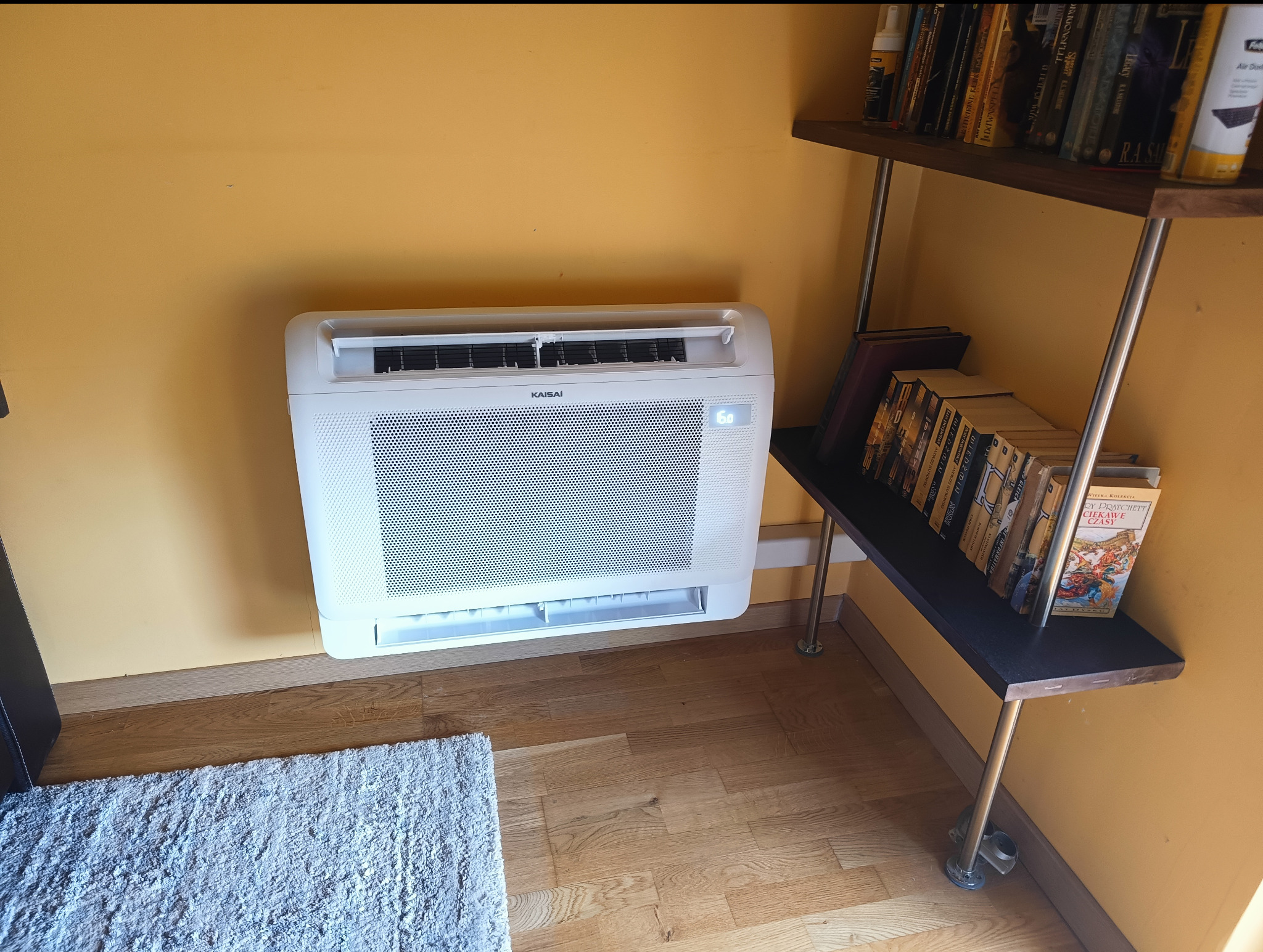 Biała klimatyzacja Kaisai na żółtej ścianie w pomieszczeniu z drewnianą podłogą i regałem na książki. Widoczny fragment dywanu.