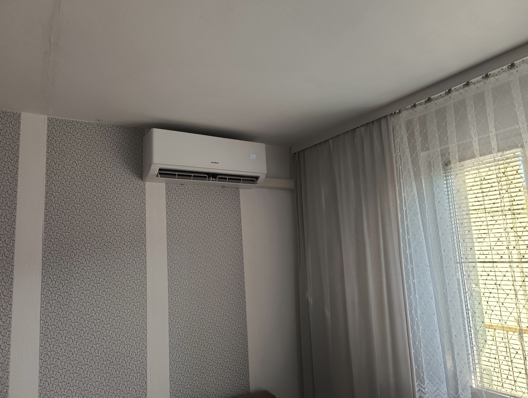 Biały klimatyzator Kaisai na ścianie w pokoju. Widoczny wyświetlacz z temperaturą 22 stopnie. Obok okno z zasłoną i żaluzjami.
