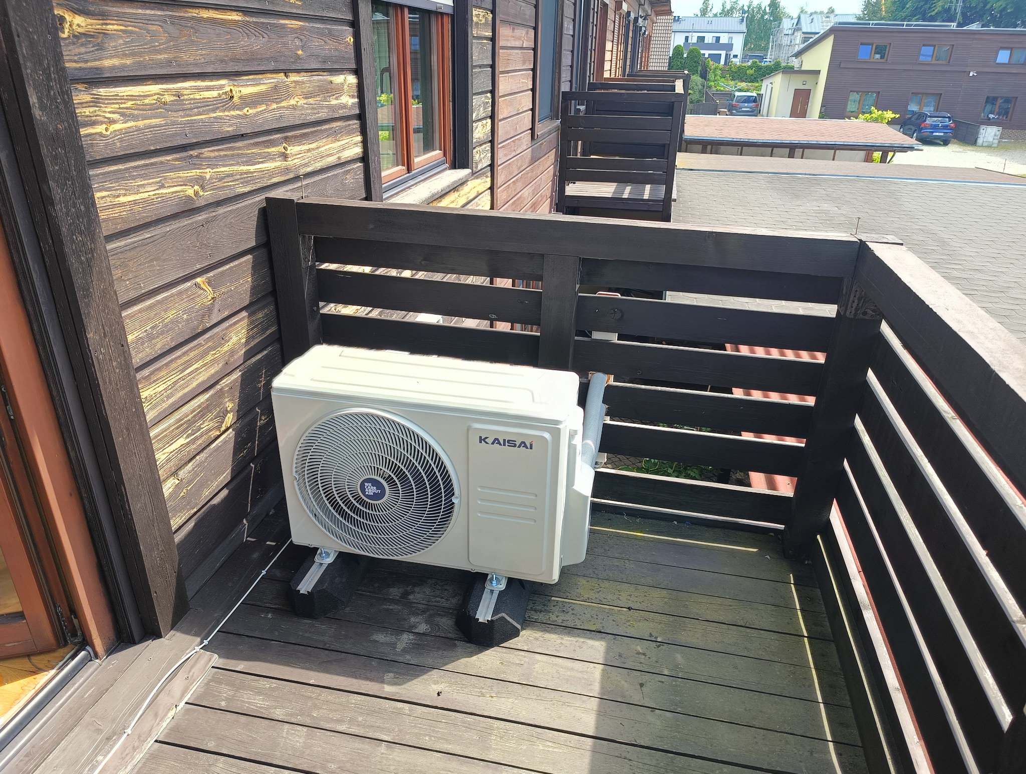 Kompaktowa klimatyzacja Kaisai zamontowana na drewnianym balkonie w budynku o elewacji z desek. Widoczne przewody i elementy montażowe.