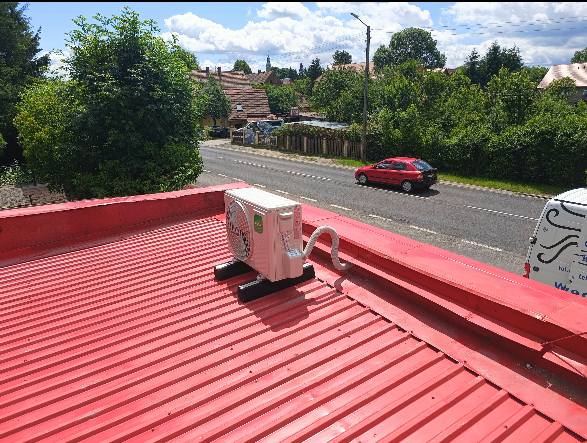 Biała jednostka zewnętrzna klimatyzacji Kaisai zamontowana na czerwonym blaszanym dachu budynku, widok na ulicę i zieleń w tle.
