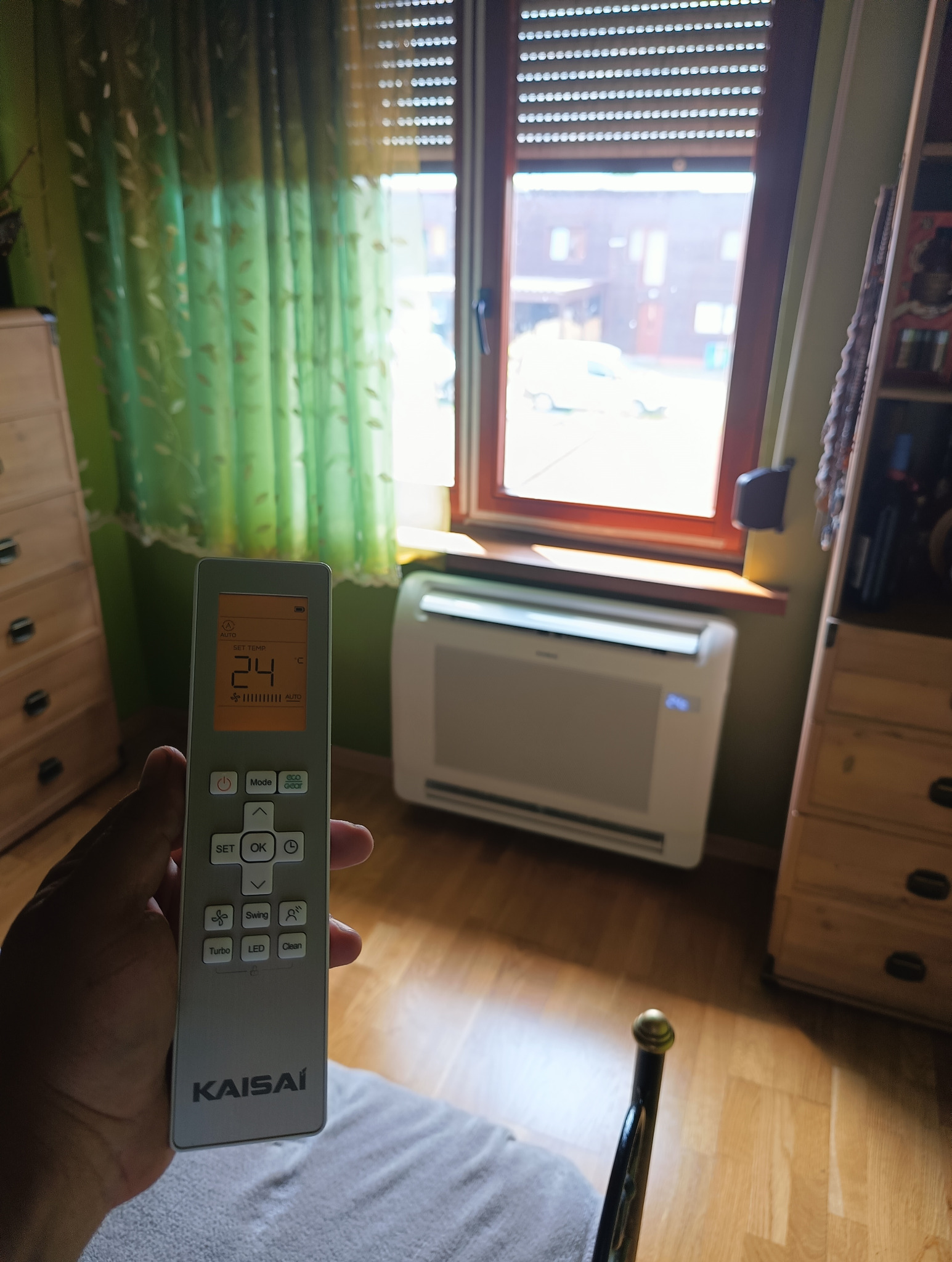 Klimatyzator Kaisaí w pokoju, sterowany pilotem ustawionym na 24 stopnie Celsjusza, z widokiem na budynek przez okno z żaluzjami.