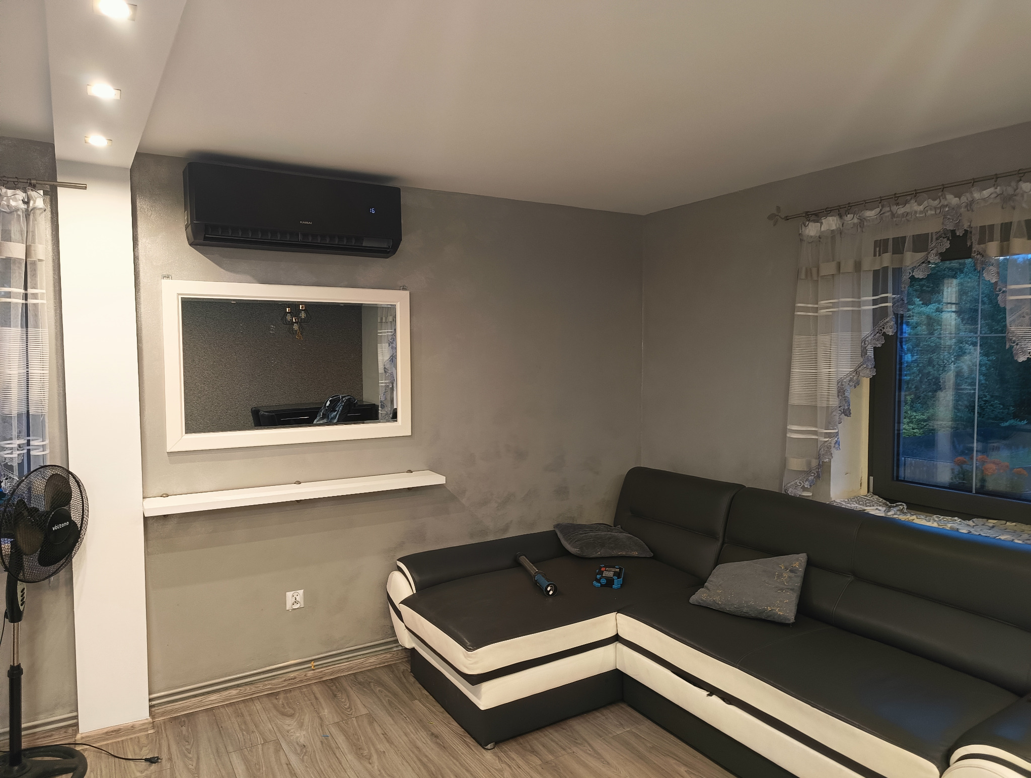 Nowoczesny klimatyzator Karsai w salonie z szarą ścianą, lustrem w białej ramie i czarno-białą sofą narożną. Widoczny wentylator w rogu pokoju.