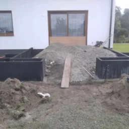 Fundamenty domu w trakcie budowy: czarne bloczki fundamentowe, g&oacute;ra żwiru, narzędzia budowlane, widok na biały budynek i trawnik. Etap prac fundamentowych.