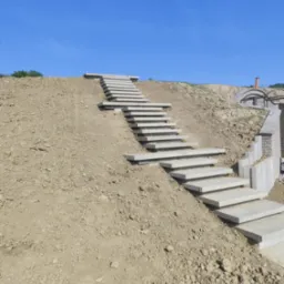 Schody z betonowych płyt prowadzące na wzgórze pod błękitnym niebem. Elementy architektury ogrodowej w tle, ziemia wokół schodów. Ujęcie z dołu.