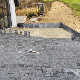 JUZBA BUDOWNICTWO Mateusz Juzba - Częściowo ułożona, szara kostka brukowa na posesji z mini koparką w tle. Widoczny pracownik przy układaniu, stosy kostki i młotek brukarski.