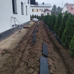 JUZBA BUDOWNICTWO Mateusz Juzba - Wykopany rów wzdłuż żywopłotu z czarnymi obrzeżami, przygotowany do montażu. Widoczne rury i przewody przy budynku. Prace ziemne w toku.